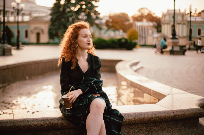 Young thoughtful red head woman sitting on fountain in city