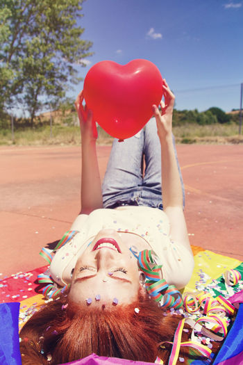 Young woman holding red heart shape balloon while lying on rainbow flag