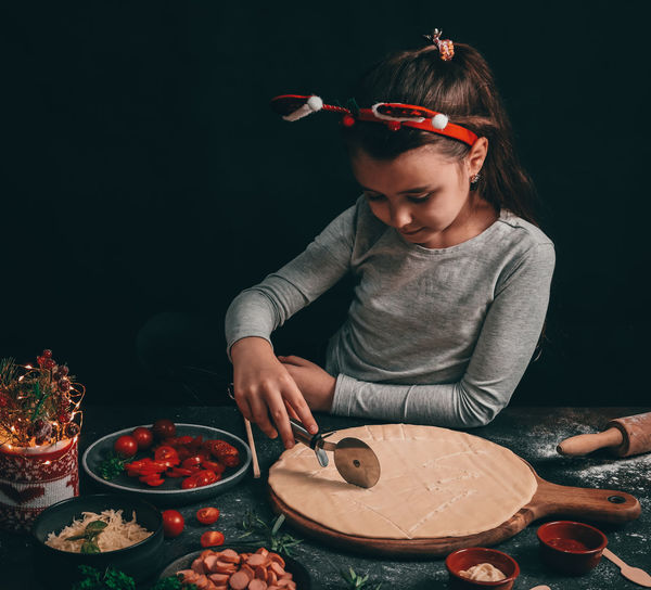 A caucasian little girl cuts a christmas tree with a round moon knife on a pizza dough.