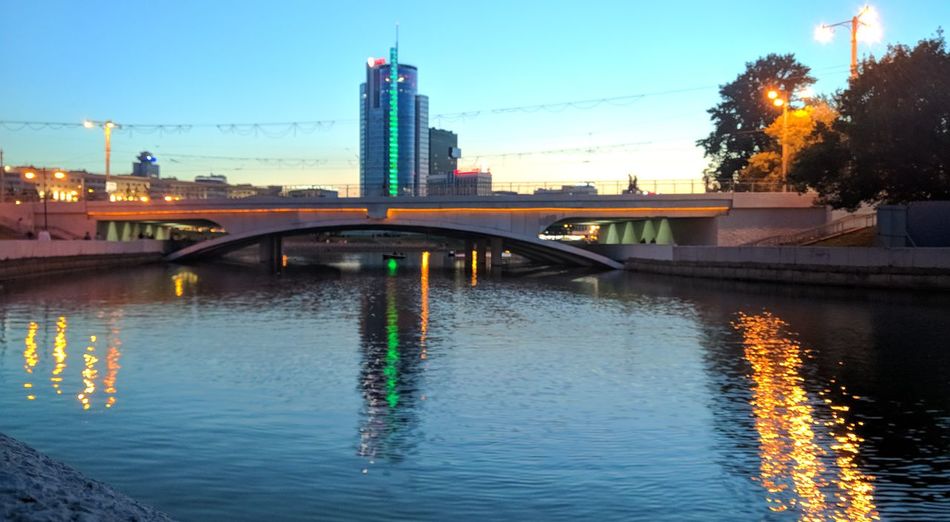 Bridge over river by illuminated buildings against sky at dusk