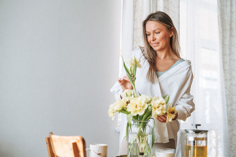 Young smiling woman in white shirt with bouquet of yellow flowers in hands near dinner table at home