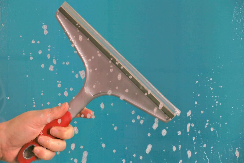 Cropped hand cleaning wet glass window with squeegee against turquoise background