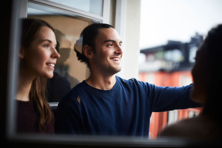 Smiling man with friends looking through window while standing in rental apartment