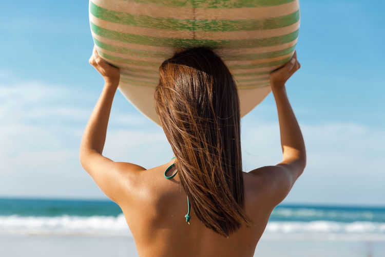 Rear view of young woman holding surfboard while standing at beach against sky