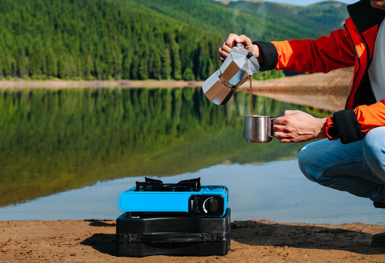 Man holding coffee drink by the camping stove against the lake