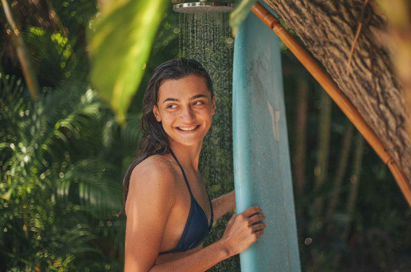 Smiling young woman with surfboard against trees