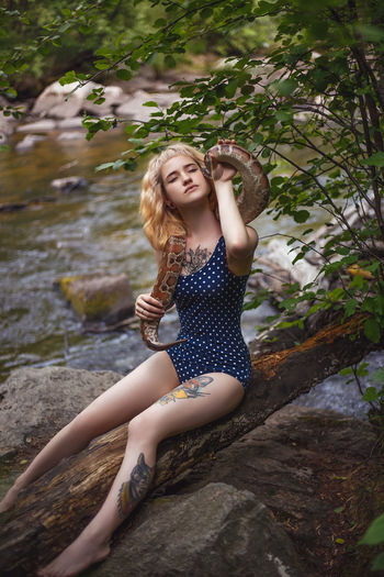 Young woman holding snake while sitting on fallen tree at forest