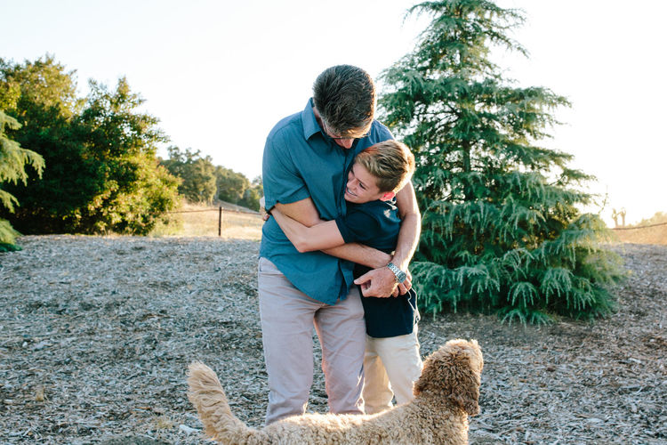 A father and teen son embrace while outside with their labradoodle dog