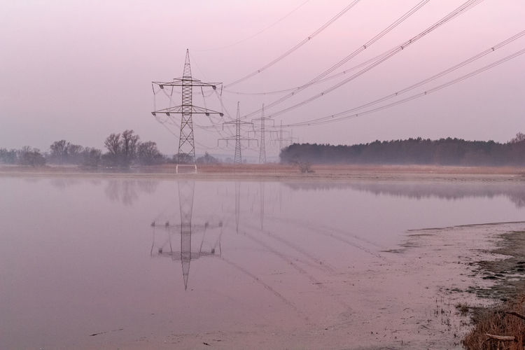 Electricity pylon by lake against sky during foggy weather