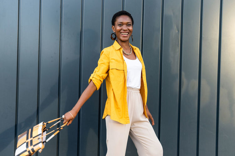 Happy adult african american female entrepreneur with short dark hair in stylish smart casual clothes standing near gray wall and looking away