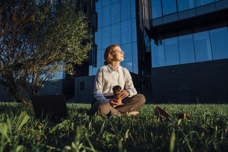 Businesswoman with disposable coffee cup sitting on grass in sunlight