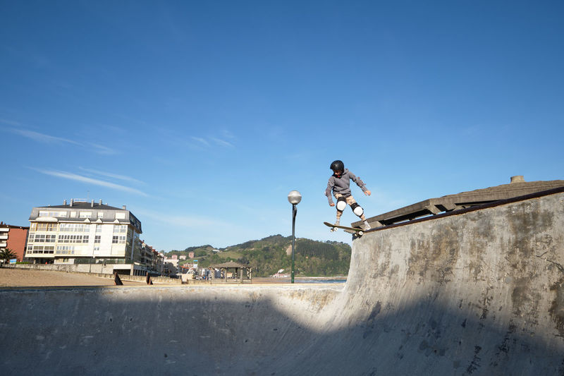 Unrecognizable teen boy in protective helmet riding skateboard in skate park on sunny day on seashore