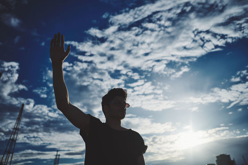 Silhouette man looking at camera while standing against sky with arm raised
