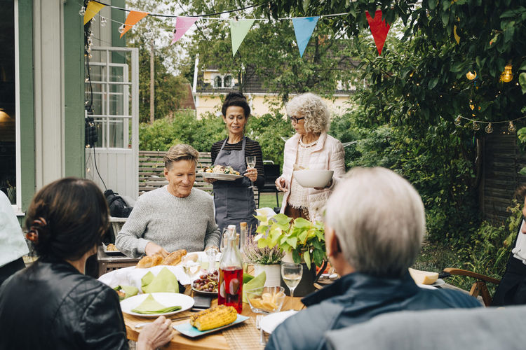 Active senior women bringing meal to friends sitting at dining table during garden party