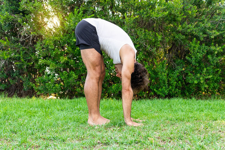 Barefoot male in activewear touching green grass while doing uttanasana pose near green bushes during yoga session in summer park