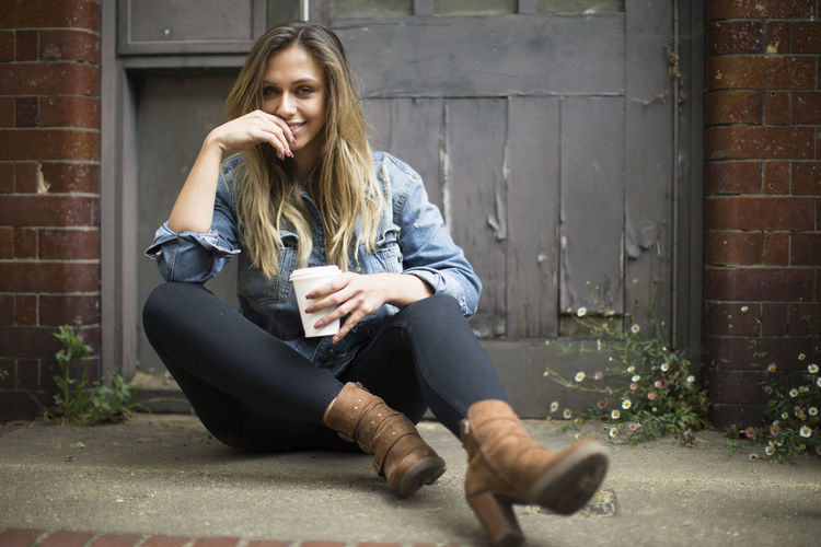 Portrait of smiling young woman with disposable cup sitting on sidewalk