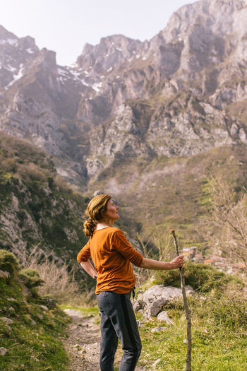 Side view of active female traveler with wooden stick standing on narrow path and admiring magnificent scenery of picos de europa mountain range with snowy rocky peaks and green slopes in spring day