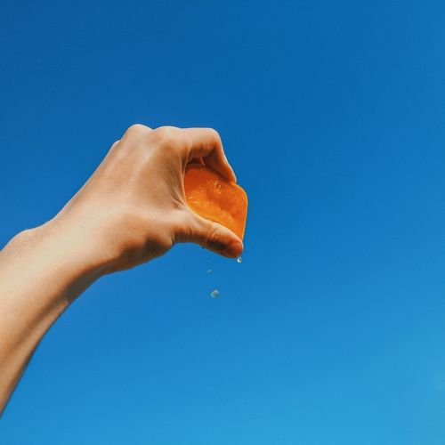Cropped hand squeezing orange against clear blue sky