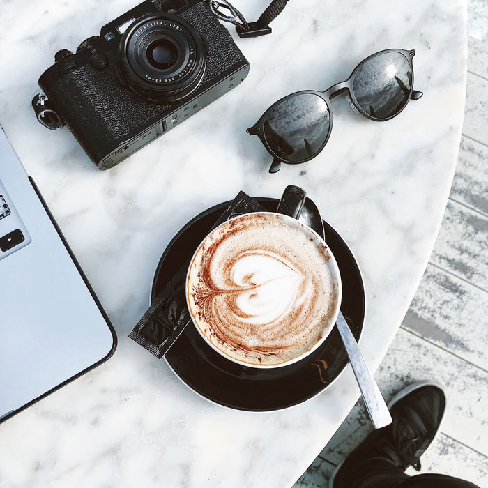 High angle point of view of coffee, laptop, camera and sunglasses flat lay on white marble table