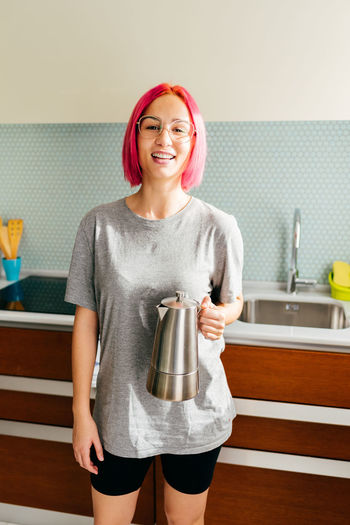 Cheerful young female with dyed hair smiling and looking at camera while preparing coffee in modern kitchen in morning