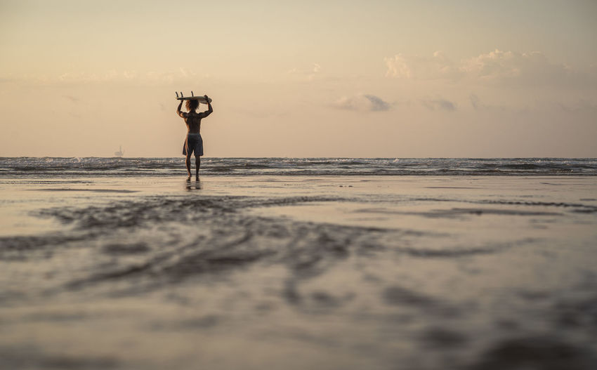 Man carrying surfboard on head while walking at seashore during sunset