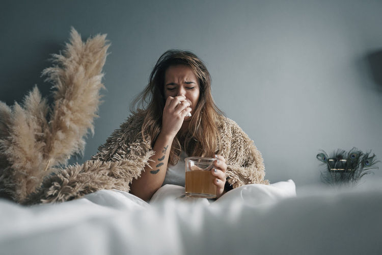 Young woman with herbal tea sitting on bed against wall