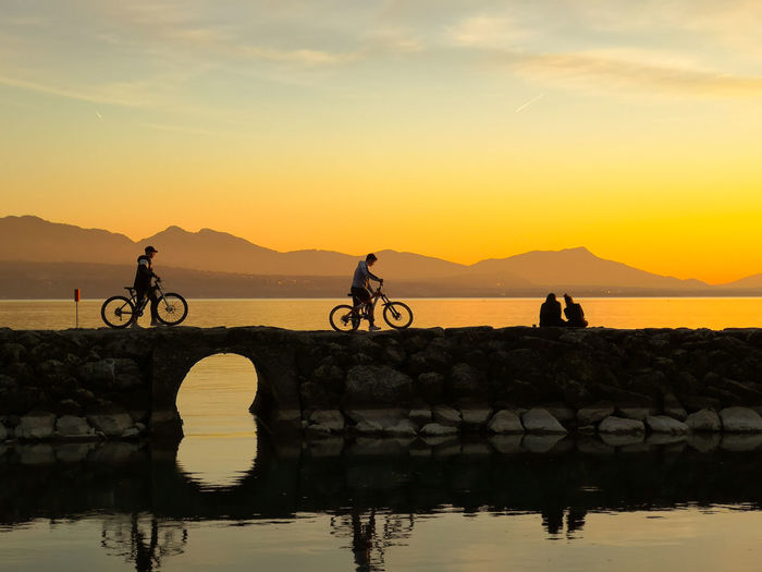 People riding bicycle by lake against sky during sunset