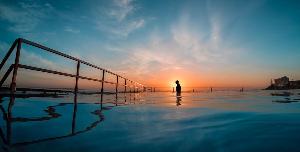 Mid distance view of silhouette man walking in sea against sky during sunset