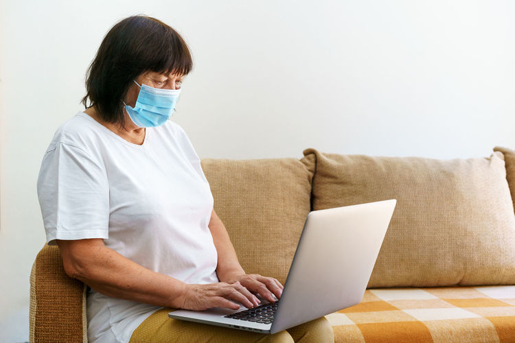 Serious woman in protective mask uses laptop, checks email on internet while