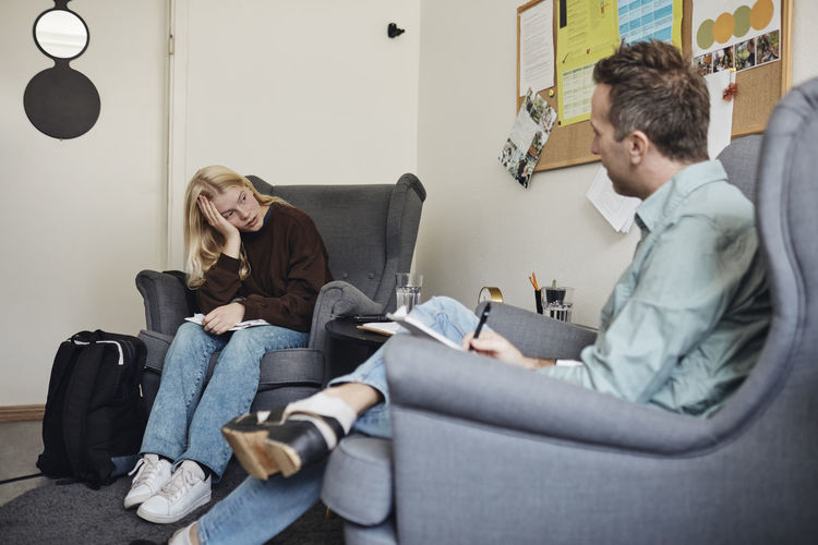 Male psychotherapist discussing with depressed female student sitting on chair in school office