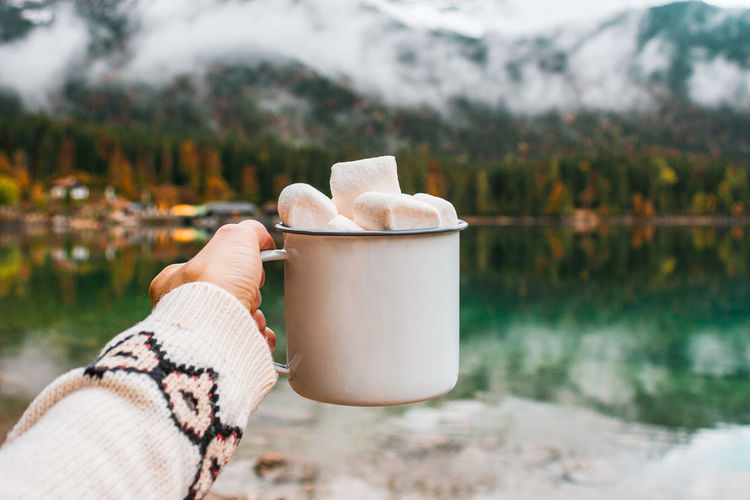 Picnic in bavarian mountains, germany. hand with mug of cacao and marshmallow on the lake background