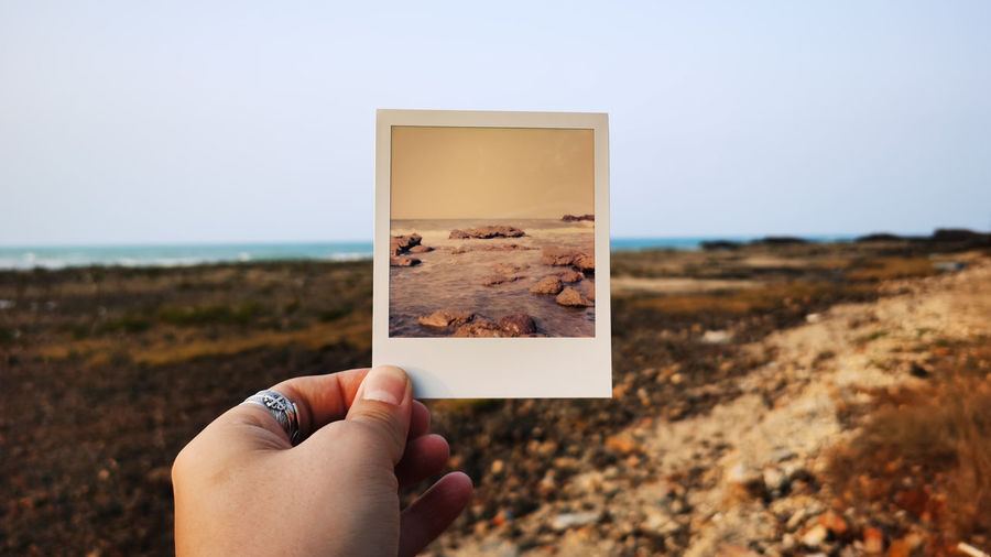 Cropped image of person holding instant print transfer on beach at sea against sky