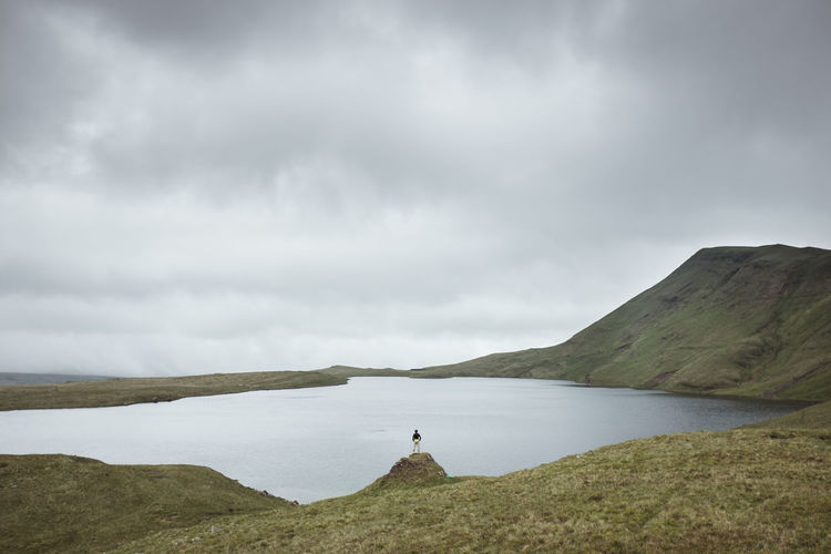 Distant view of man standing by lake against cloudy sky