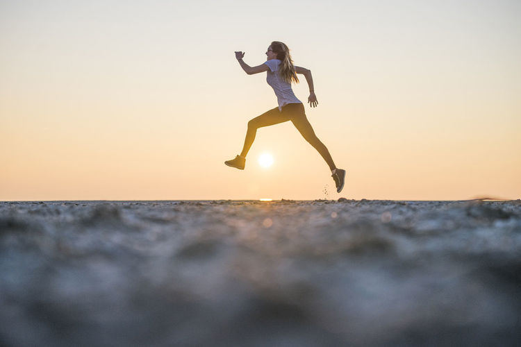 Low angle view of young woman jumping on beach against sky during sunset