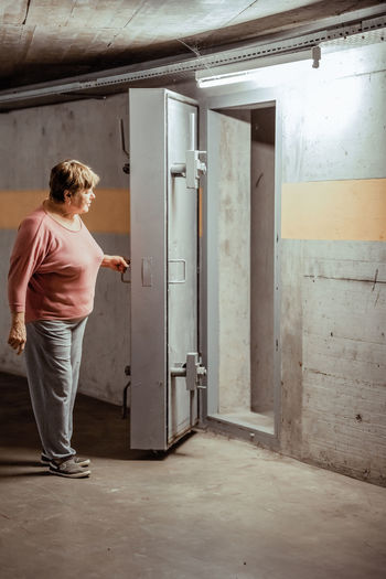 Senior woman entering nuclear fallout shelter built in basement of building opening thick metal door