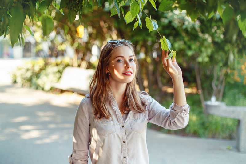 Portrait of young woman touching tree while standing in park