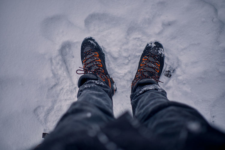First person view on a man's legs with a black jeans and black and orange hiking boots on snow.