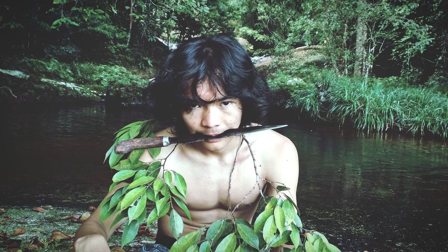 Portrait of shirtless young man carrying knife in mouth by river at forest
