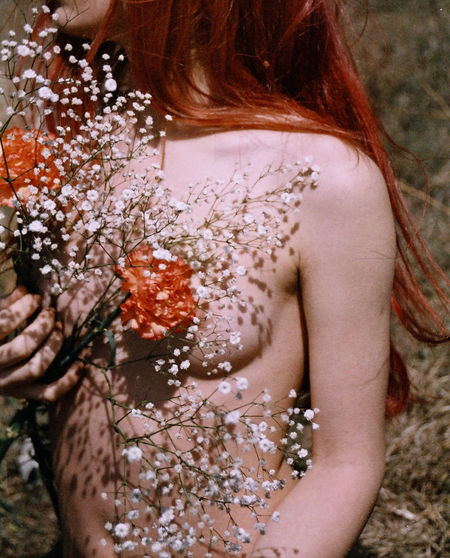 Midsection of shirtless woman covering breast with flowers while standing outdoors