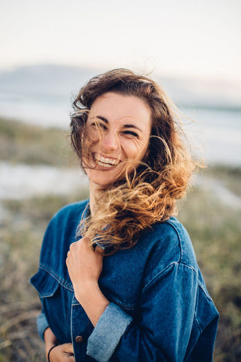 Portrait of cheerful young woman standing at beach against clear sky during sunset
