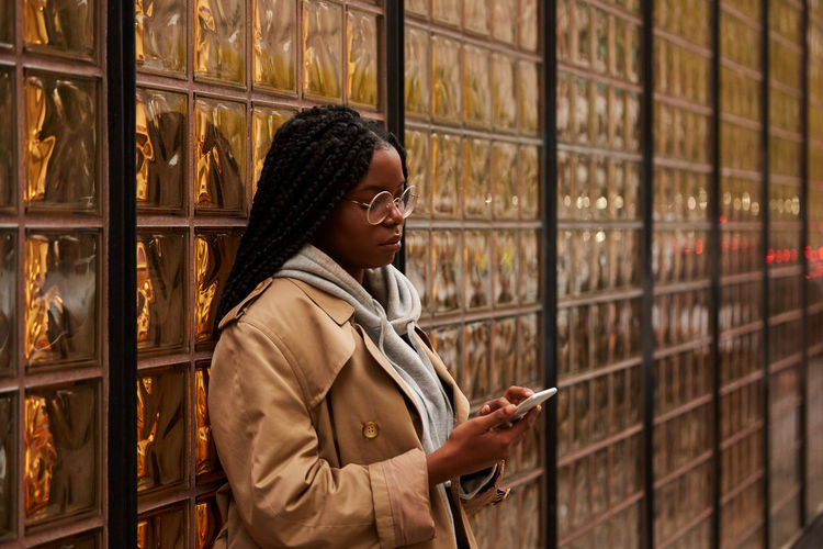 Confident black female with braids in casual outfit looking away and browsing smartphone while standing near glass wall