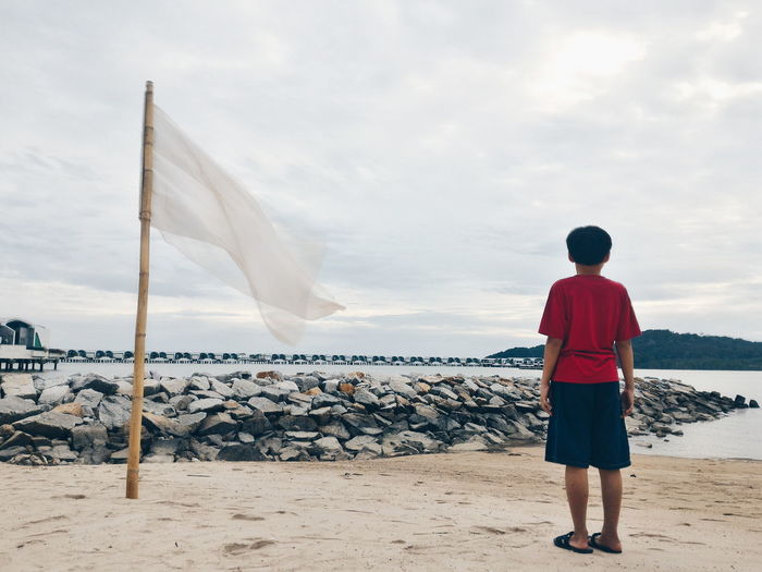 Full length rear view of boy standing by white flag at beach against sky