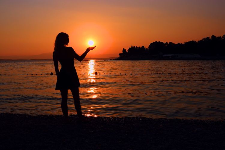 Optical illusion of silhouette woman holding sun while standing at beach against sky during sunset