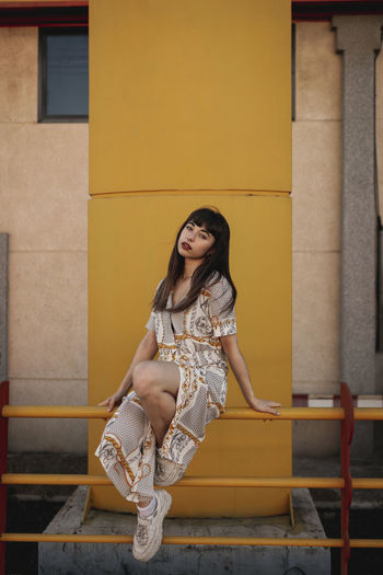 Full body of young self esteem ethnic female with long dark hair in stylish outfit sitting on metal railing near modern building and looking at camera