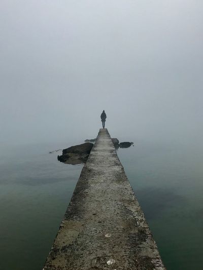 Distant view of woman standing on pier by sea during foggy weather