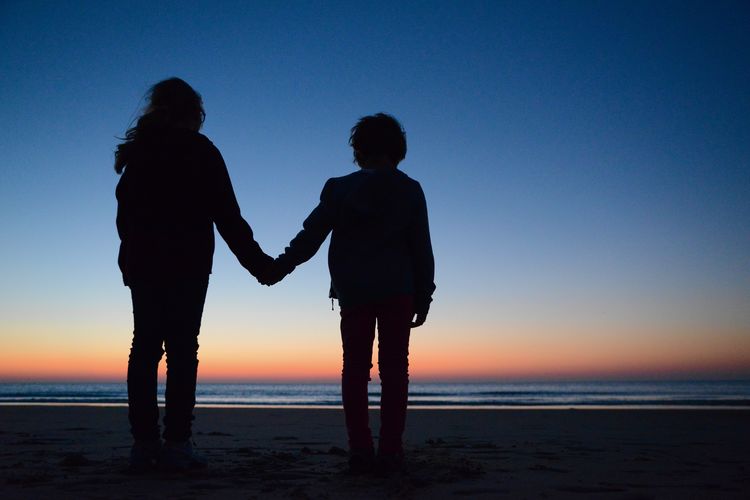 Silhouette siblings holding hands while standing at beach against sky during sunset