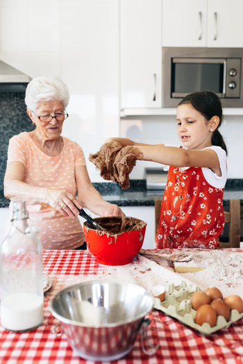 Girl with dough on hands against cheerful grandma at table with bowl during cooking process at home