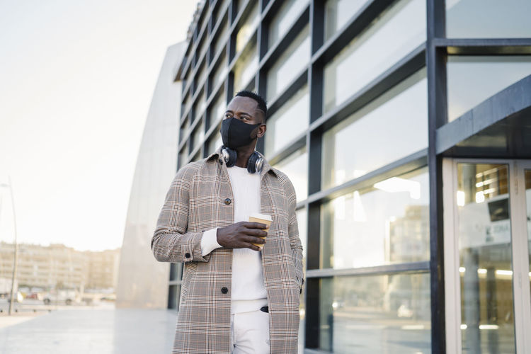 Fashionable man wearing protective face mask holding coffee cup against building