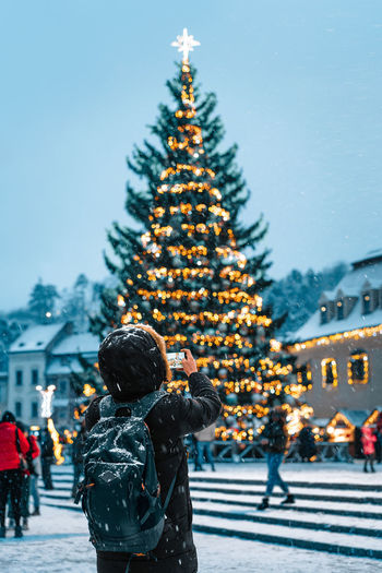 Rear view of person in warm clothing photographing christmas market and tree in snowy winter night