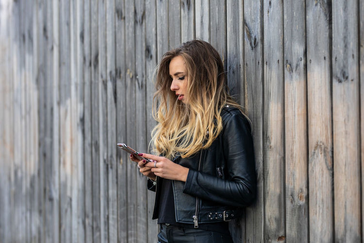 Young woman with blond hair using phone by wooden fence